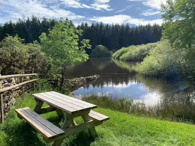 a wooden picnic table sitting next to a lake at The Nook at Pentregaer Ucha, with tennis court & lake. in Oswestry