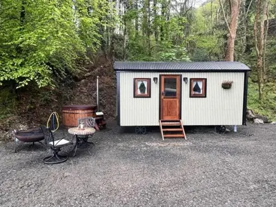 a small white shed with a table and a chair at Romantic Shepherds Hut with Hot Tub in The Heart of Snowdonia in Dolgellau